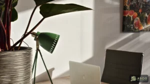 Op een houten bureau staat een geopende laptop naast een groene bureaulamp en een grote kamerplant. Zonlicht valt over de werkplek en een donkere bureaustoel.