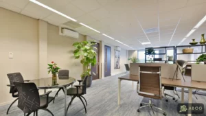 Een lichte kantoorruimte met links een ronde tafel met zwarte stoelen en rechts werkplekken met bureaus, bureaustoelen en kamerplanten.