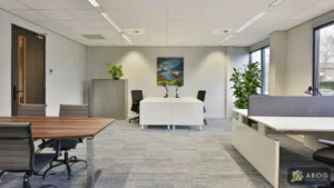 Een lichte kantoorruimte met een houten vergadertafel en witte bureaus met bureaustoelen op een grijze vloerbedekking. Aan de achterwand hangt een schilderij.