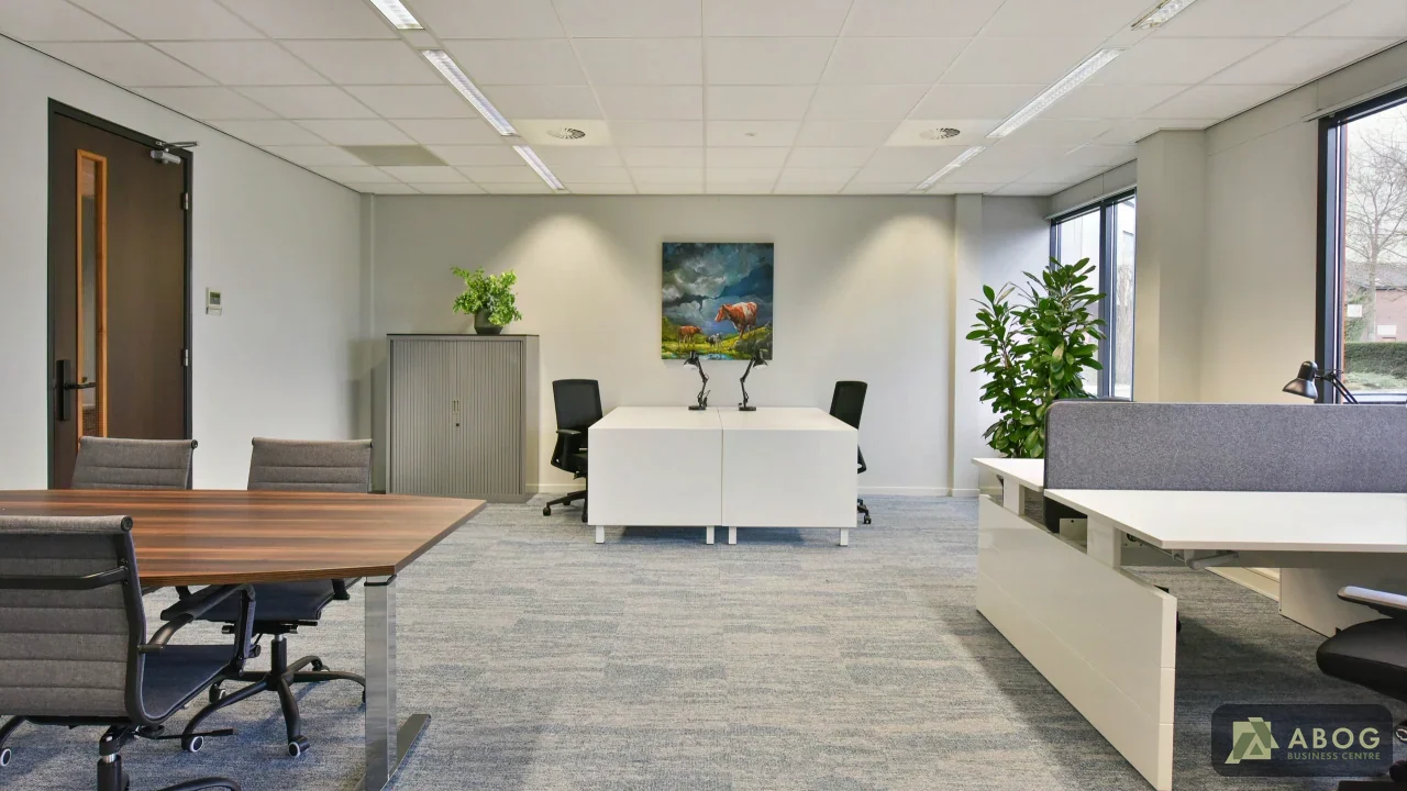 Een lichte kantoorruimte met een houten vergadertafel en witte bureaus met bureaustoelen op een grijze vloerbedekking. Aan de achterwand hangt een schilderij.