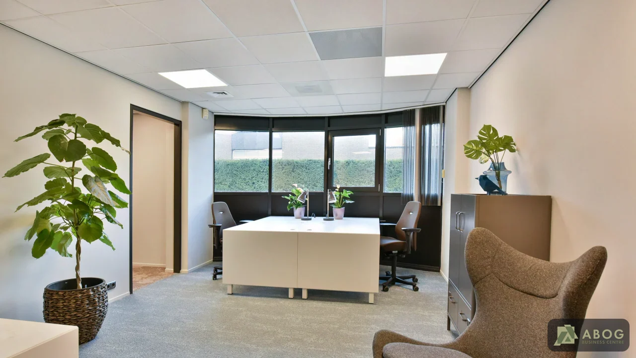 Een lichte kantoorruimte met een wit bureau, bureaustoelen, een fauteuil en grote kamerplanten. Grote ramen aan de achterzijde laten daglicht binnen.