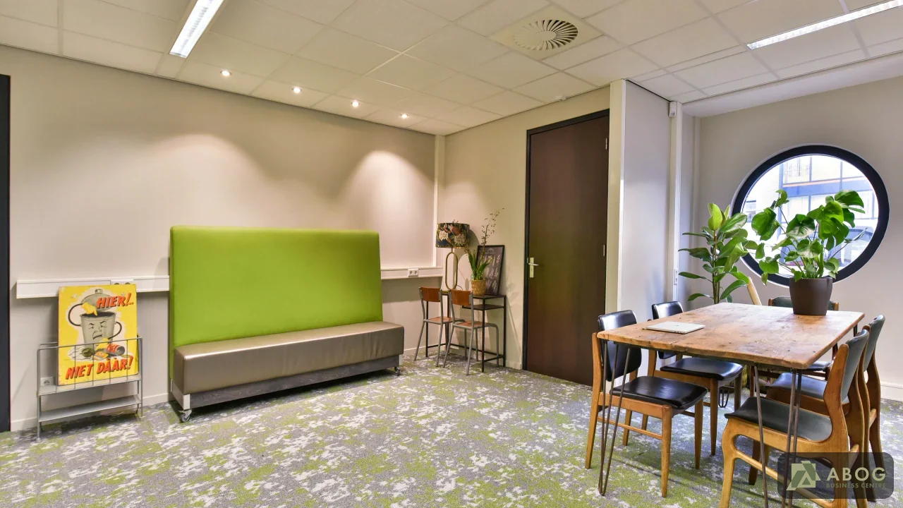Een kantoorruimte met een houten vergadertafel met stoelen, een hoge felgroene bank en een rond raam. Op de vloer ligt een groen met grijs gemêleerd tapijt.