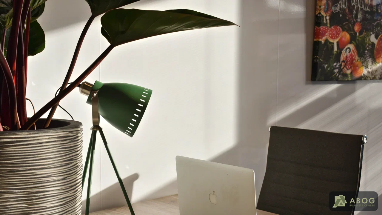 Op een houten bureau staat een geopende laptop naast een groene bureaulamp en een grote kamerplant. Zonlicht valt over de werkplek en een donkere bureaustoel.