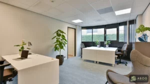 Een lichte kantoorruimte met witte bureaus, bureaustoelen, kamerplanten en een fauteuil. Grote ramen aan de achterzijde laten daglicht binnen.